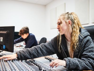A girl with blonde hair and a grey jumper sits at a mixing desk. Her right hand is operating the controls